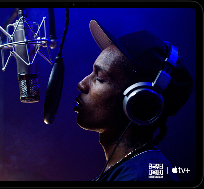 가로 방향으로 놓인 스페이스 블랙 색상 iPad Pro의 앞면. 둥근 형태의 모서리, 블랙 디스플레이 베젤이 보이고 화면에는 Apple TV+ 오리지널 영화 '천국부터 지옥까지' - Highest 2 Lowest에 등장하는 Yung Felon 역의 에이셉 라키가 마이크 앞에서 랩을 하고 있는 모습이 재생되고 있습니다.